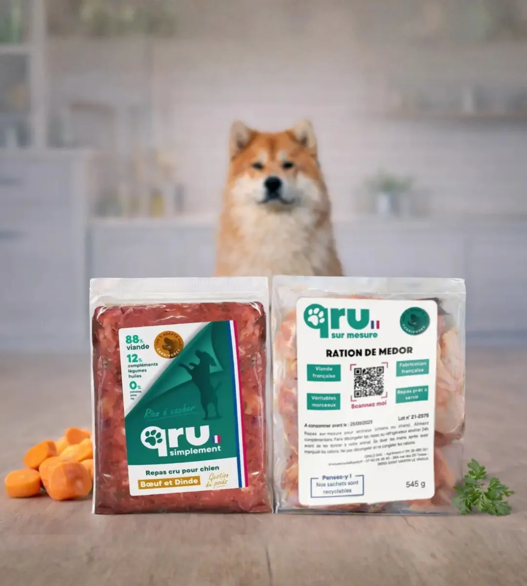 Deux paquets de repas BARF pour chien, un BARF haché et un BARF en morceaux, posés sur une table avec des légumes, un chien en arrière-plan dans une cuisine.